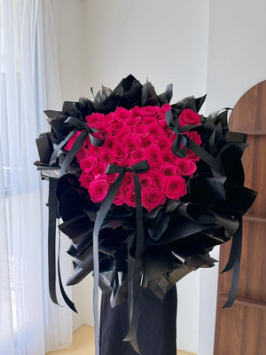 A large bouquet of vibrant 52 hot pink roses for proposal wrapped in black paper and tied with black ribbons, displayed indoors against a white wall and curtain.