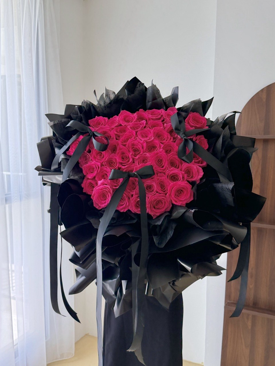 A large bouquet of vibrant 52 hot pink roses for proposal wrapped in black paper and tied with black ribbons, displayed indoors against a white wall and curtain.