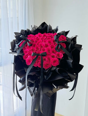 A large bouquet of vibrant hot pink roses wrapped in black paper with black ribbons, displayed on a stand in front of a white curtain, perfect for proposal.