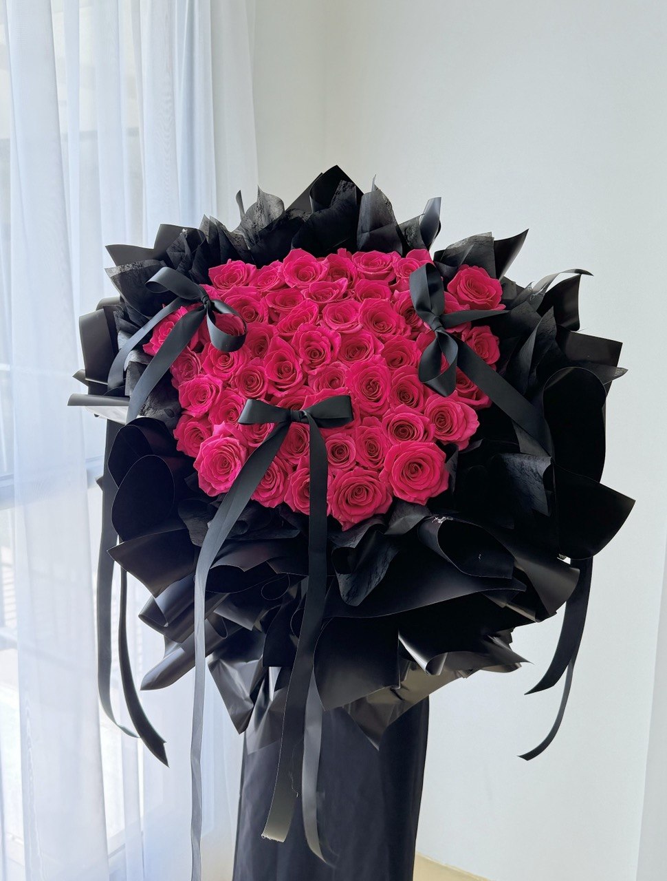 A large bouquet of vibrant hot pink roses wrapped in black paper with black ribbons, displayed on a stand in front of a white curtain, perfect for proposal.