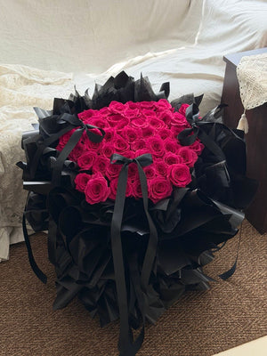 A large bouquet 52 hot pink roses for proposal wrapped in black paper and tied with black ribbons, placed on a brown carpeted floor in front of a white bed.