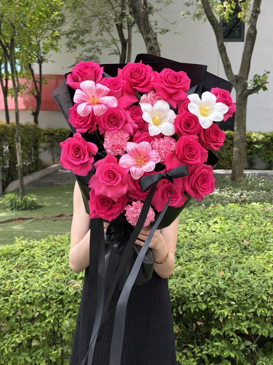 A person outdoors holding a large bouquet of hot pink roses, dyed tulip, white tulip and dyed carnation wrapped in black paper with black ribbons. The background shows green shrubs and trees.
