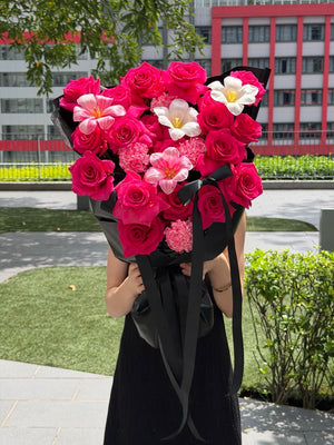 A person in a black dress holds a large bouquet of pink roses, dyed tulip, white tulip and dyed carnation wrapped in black paper with long ribbons, outdoors in front of a modern building and greenery.