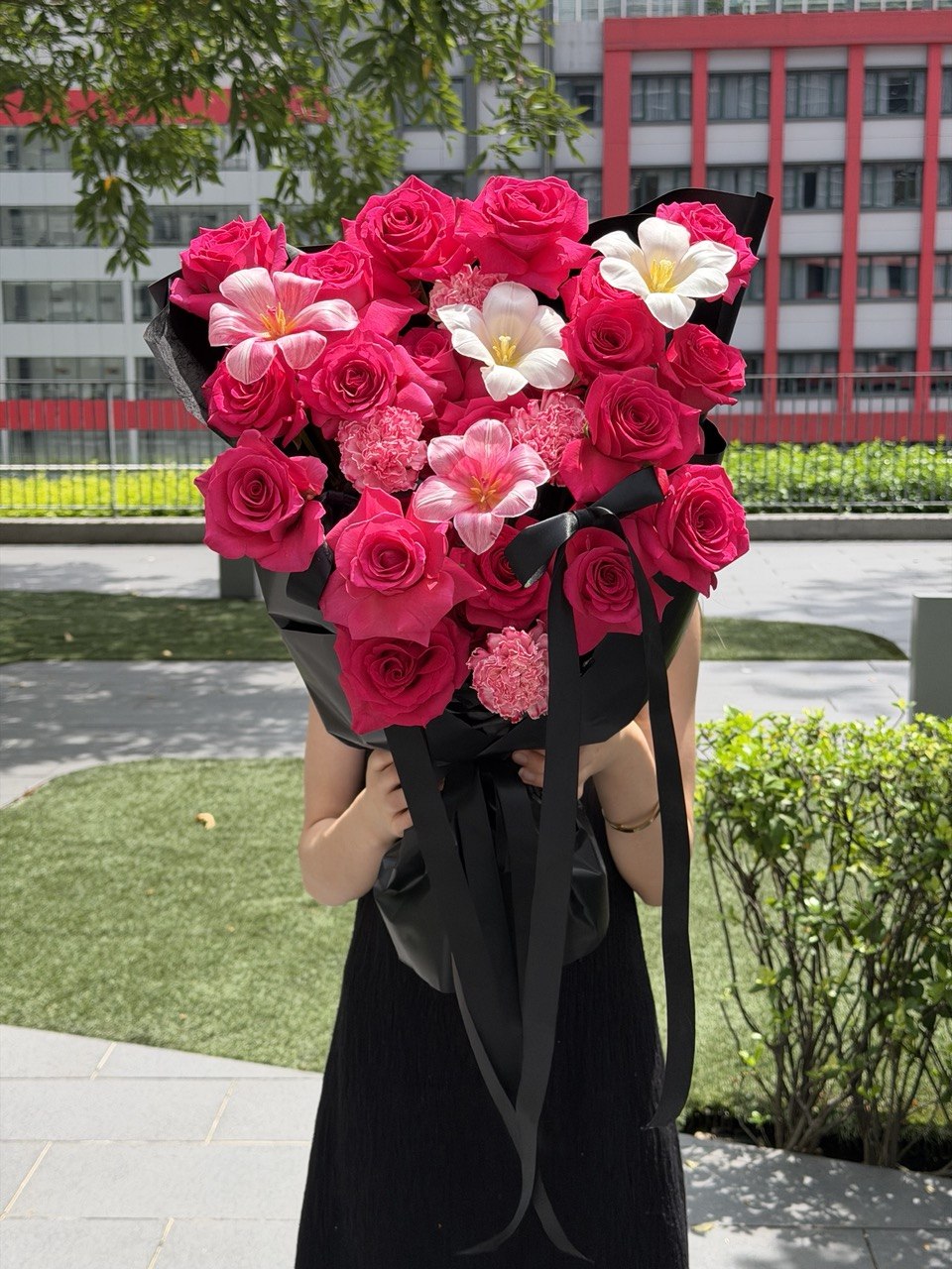 A person in a black dress holds a large bouquet of pink roses, dyed tulip, white tulip and dyed carnation wrapped in black paper with long ribbons, outdoors in front of a modern building and greenery.