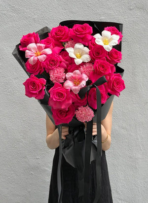 A person holding a large bouquet of vibrant pink roses, dyed tulip, white tulip and dyed carnation wrapped in black paper, against a textured gray wall.
