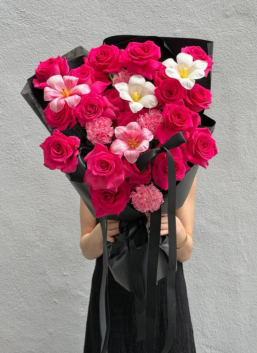 A person holding a large bouquet of vibrant pink roses, dyed tulip, white tulip and dyed carnation wrapped in black paper, against a textured gray wall.