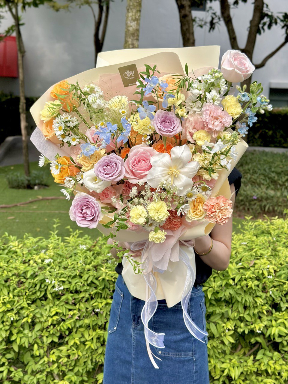 A lush, garden-style bouquet with layered pastel blooms and delicate wrapping, held by a woman in a navy top and denim skirt.