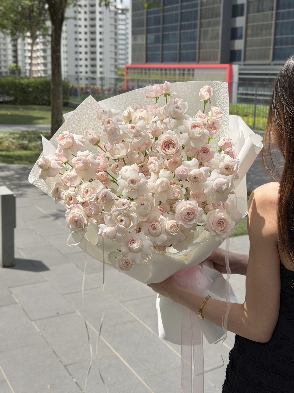 Person holding a large bouquet of pink kiss of fairy rose spray outdoors with buildings in the background