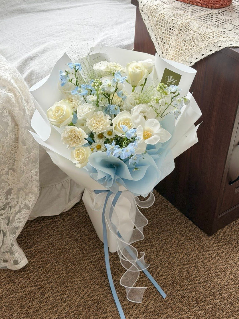 A bouquet of white and light blue flowers wrapped in white paper with a blue ribbon, placed on a carpeted floor next to a bed and bedside table.