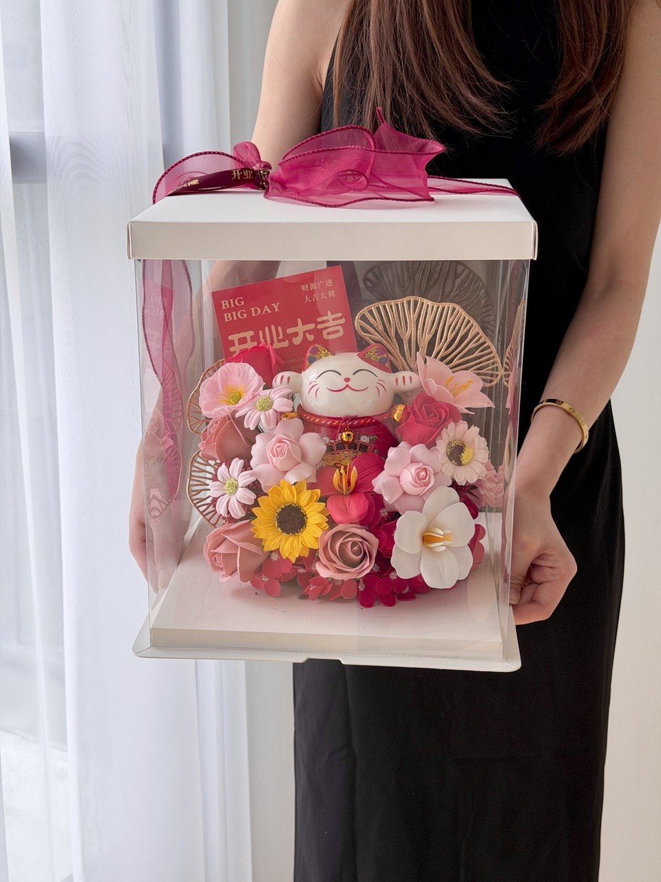 A person holds a clear square gift box with a white lid and bright pink ribbon. Inside the box is a decorative arrangement featuring a red lucky cat figurine, pink and red flowers, a yellow sunflower, and gold fan-shaped accents perfect for grand opening lucky cat gift.