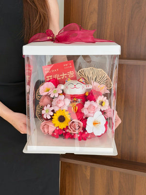 A person holds a clear acrylic gift box with a white lid and a bright pink ribbon. The box contains a decorative arrangement of pink and white flowers, a yellow sunflower, a red card, and a red lucky cat figurine, set against a wooden background.