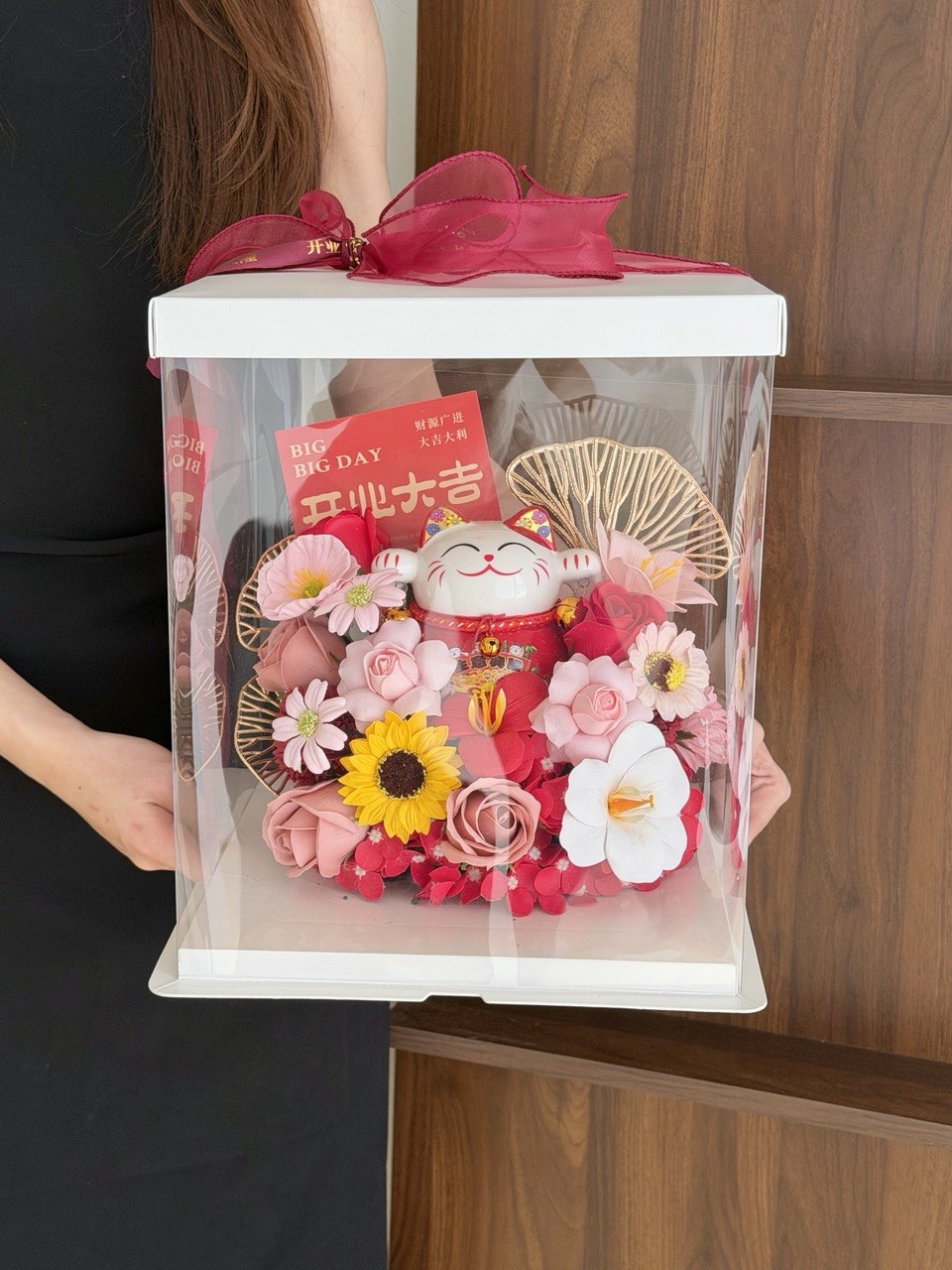 A person holds a clear acrylic gift box with a white lid and a bright pink ribbon. The box contains a decorative arrangement of pink and white flowers, a yellow sunflower, a red card, and a red lucky cat figurine, set against a wooden background.