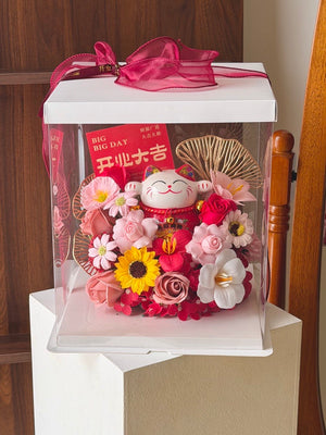 A decorative gift box containing a red maneki-neko figurine, pink and red artificial flowers, a yellow sunflower, and gold fan-shaped ornaments, displayed on a pedestal in an indoor setting.
