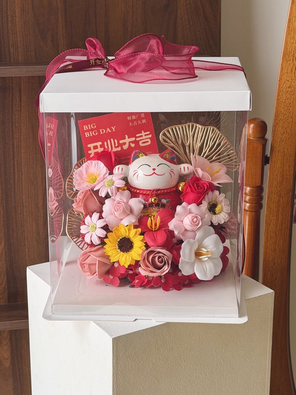 A decorative gift box containing a red maneki-neko figurine, pink and red artificial flowers, a yellow sunflower, and gold fan-shaped ornaments, displayed on a pedestal in an indoor setting.
