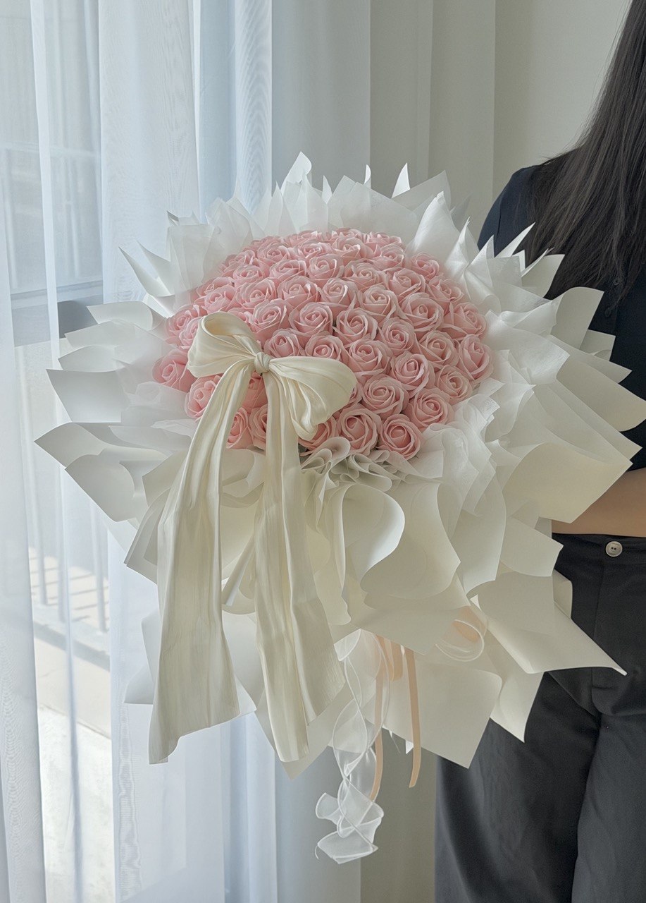 A large bouquet of 52 pink roses soap flower wrapped in white wrapper with a cream satin ribbon bow, held by a person standing in front of a window with sheer curtains, perfect bouquet for proposal.