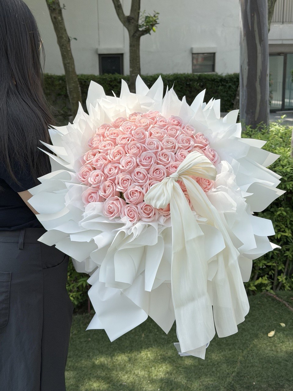 A large round-shaped bouquet of 52 pink roses soap flower wrapped in white paper and tied with a white ribbon, held by a person standing outdoors on grass with trees and a building in the background, perfect for proposal gift.