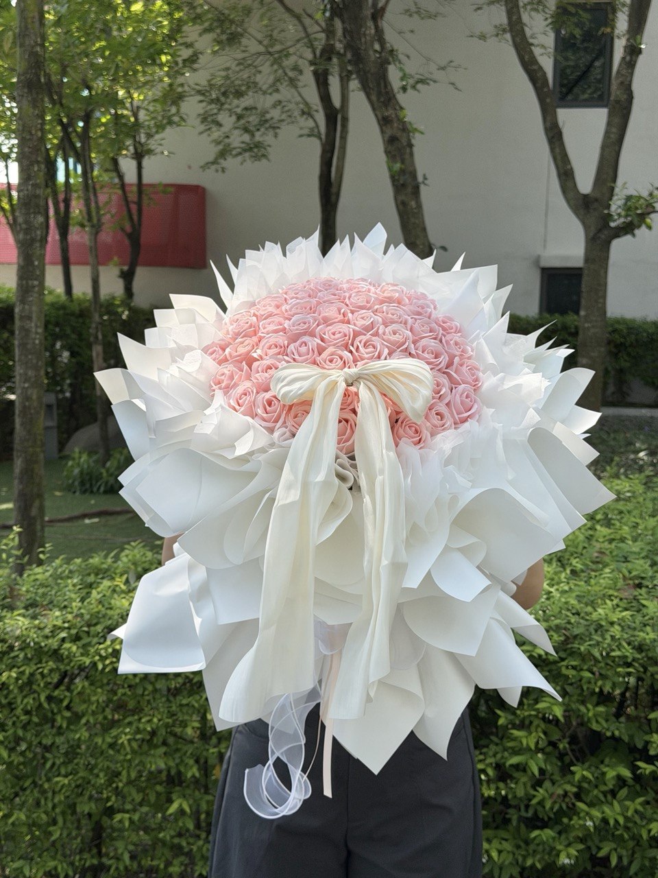 A large bouquet of 52 pink roses wrapped in white paper with a long white ribbon bow, held outdoors in front of green hedges and trees.