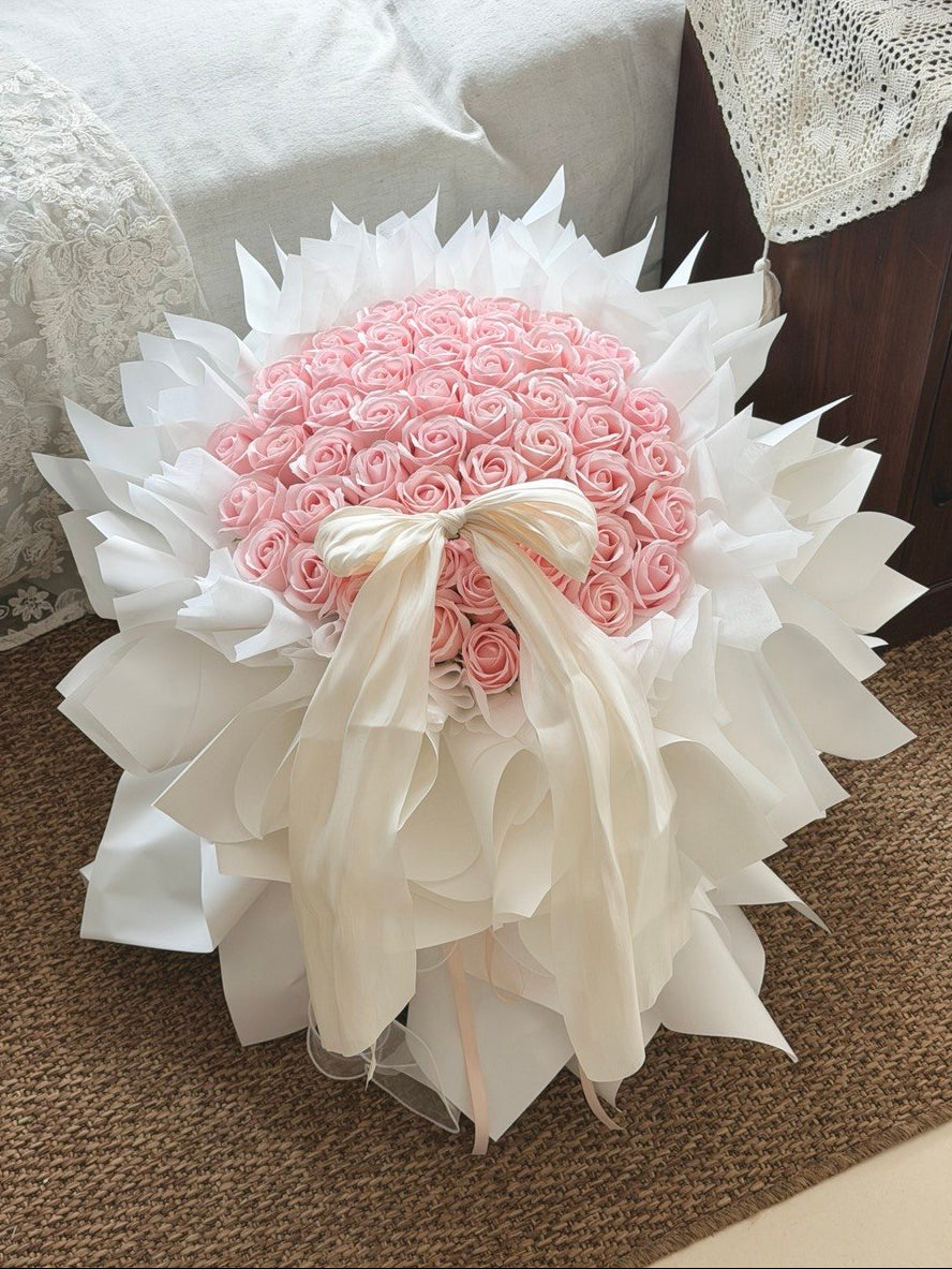 A large bouquet of 52 pink roses soap flower wrapped in white paper and tied with a cream ribbon, placed on a brown carpeted floor next to a bed with white bedding, perfect for proposal.