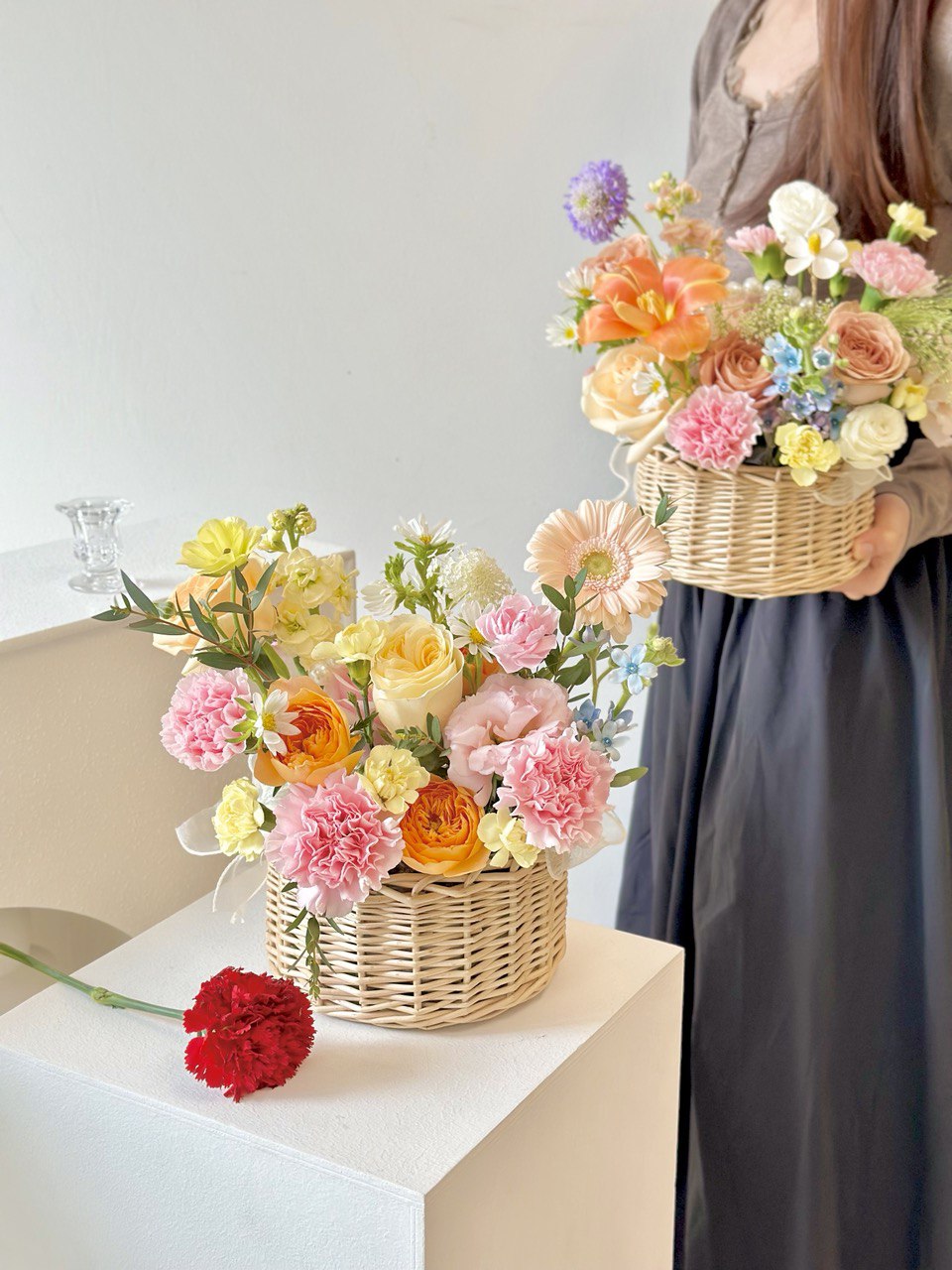 A woman wearing a brown top holds a flower basket, while another basket rests on a table, suitable for any occasion.