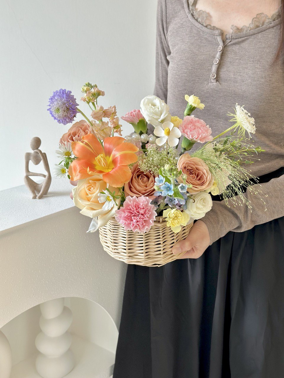A woman in a brown top carries a basket of flowers, with an additional basket on the table, ideal for various occasions.