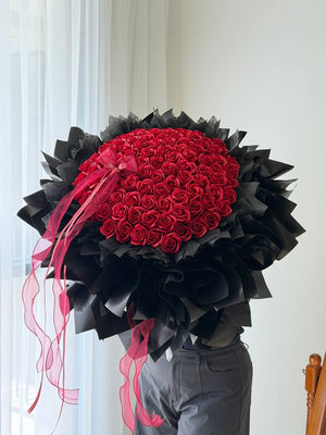 A large round 99 red roses soap flower bouquet wrapped in black paper with long pink ribbon tails, held by a person wearing gray pants in a bright indoor setting.