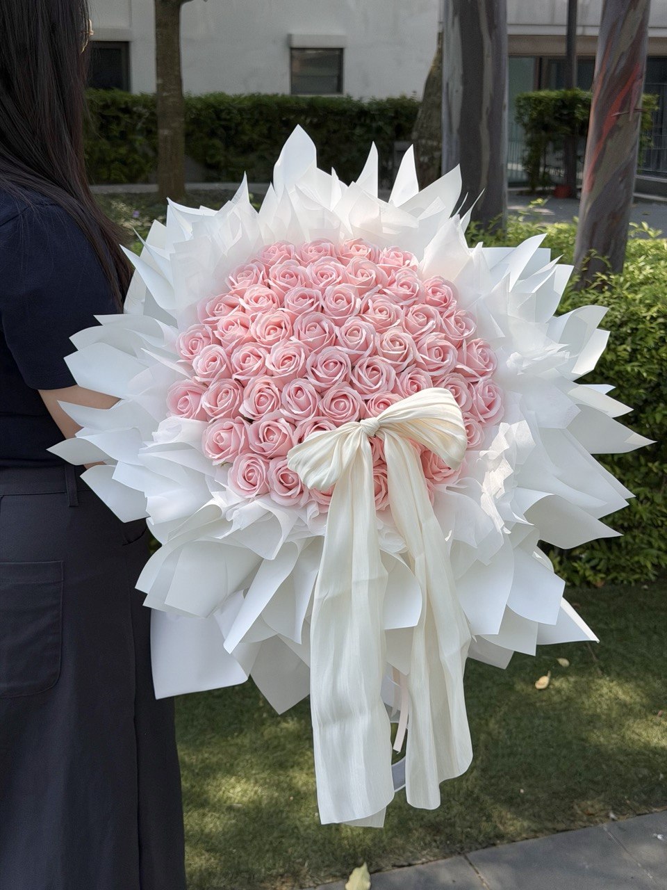 A large round-shaped bouquet of 52 pink roses soap flower wrapped in white paper and tied with a white ribbon, held by a person standing outdoors on grass with trees and a building in the background, perfect for proposal gift.