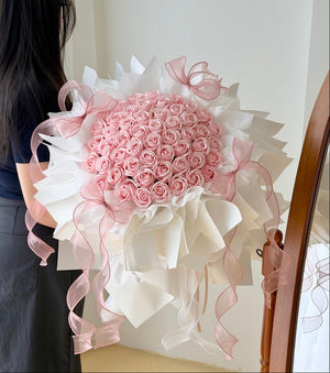 A large round bouquet of pink roses soap flower wrapped in white wrapper with long pink ribbon bows and trailing ribbons, held by a person whose back and side are visible against an indoor wall.