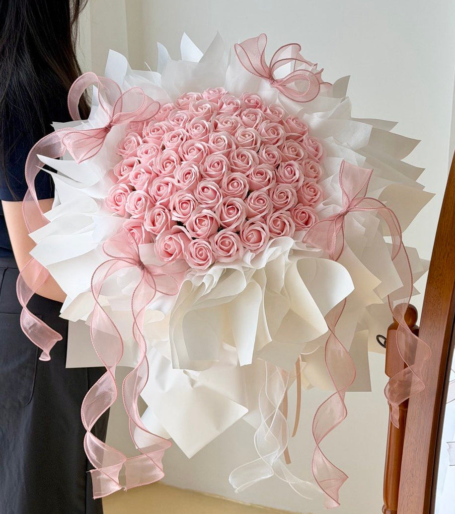 A large round bouquet of pink roses soap flower wrapped in white wrapper with long pink ribbon bows and trailing ribbons, held by a person whose back and side are visible against an indoor wall.