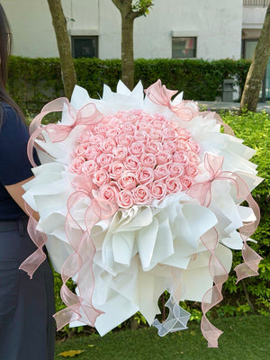 A large round-shaped bouquet of pink roses soap flower wrapped in white paper with pink ribbon bows, held by a person outdoors in front of green hedges and trees.