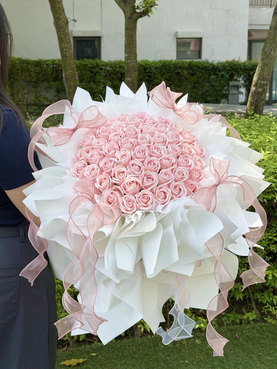 A large round-shaped bouquet of pink roses soap flower wrapped in white paper with pink ribbon bows, held by a person outdoors in front of green hedges and trees.