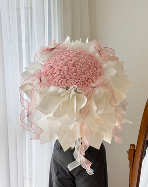 A large round bouquet of pink roses soap flower wrapped in white paper with sheer pink ribbon bows, held in front of a person wearing black pants in an indoor setting.