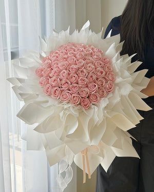 A large bouquet of pink roses soap flower wrapped in white paper, held by a person standing in front of a window with sheer curtains.