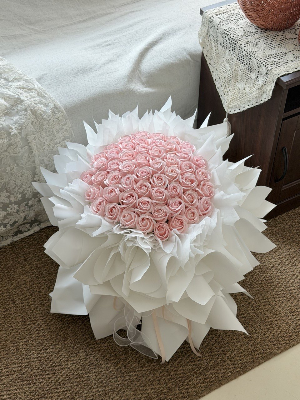 A large round bouquet of pink roses soap flower wrapped in white paper, placed on a brown carpeted floor next to a bed and bedside table.