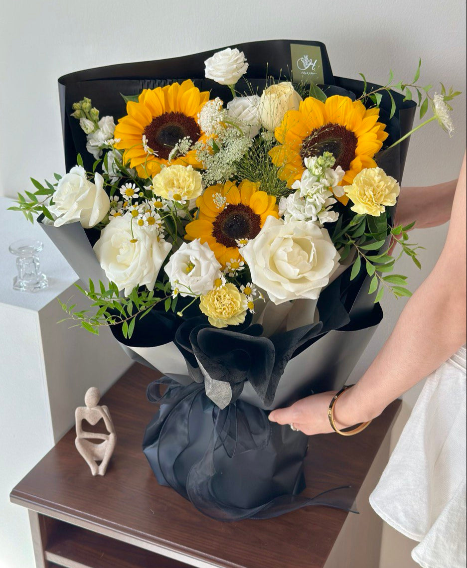 Graduation bouquet featuring vibrant sunflowers, white roses, and yellow carnations wrapped in elegant black paper.