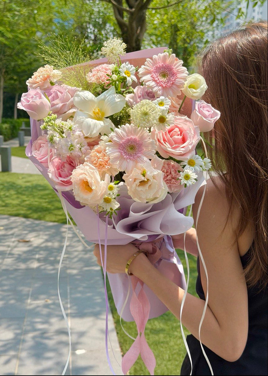 A woman outdoors holds a large bouquet of pastel pink, peach, white, and light green flowers wrapped in light purple paper with long ribbon tails. Trees, grass, and a paved walkway are visible in the background.