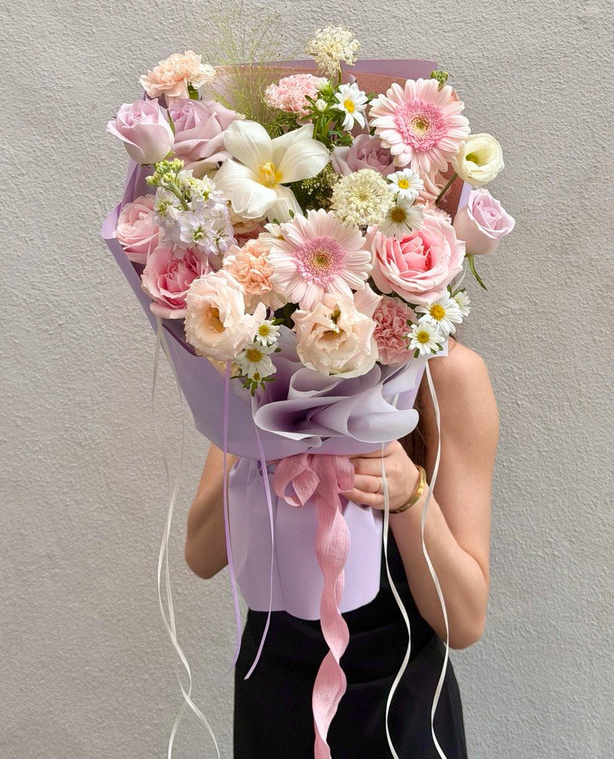 A person holding a large, beautifully arranged flower bouquet with pink roses, white lilies, and daisies wrapped in light purple paper with pink ribbons, set against a neutral gray wall.