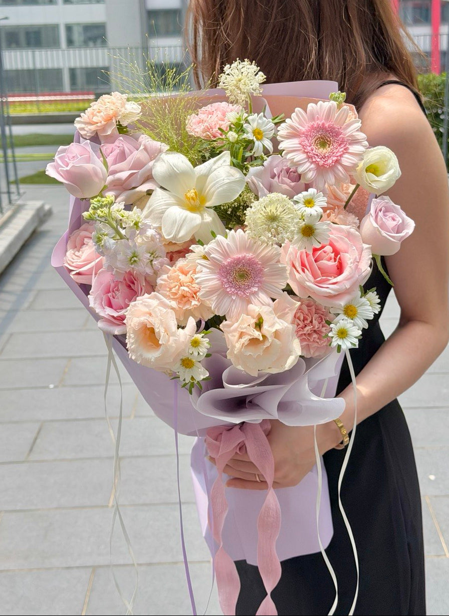 A woman holding a large, vibrant flower bouquet with pink, peach, white, and yellow flowers wrapped in light purple paper, set against an outdoor paved background.