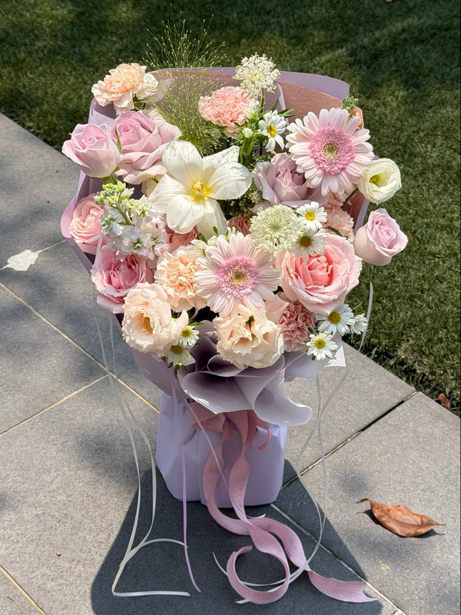 A elegant bouquet of pink, peach, white, and light purple flowers wrapped in a matching light purple paper sleeve with a long pink ribbon, placed on a gray stone path with green grass in the background.