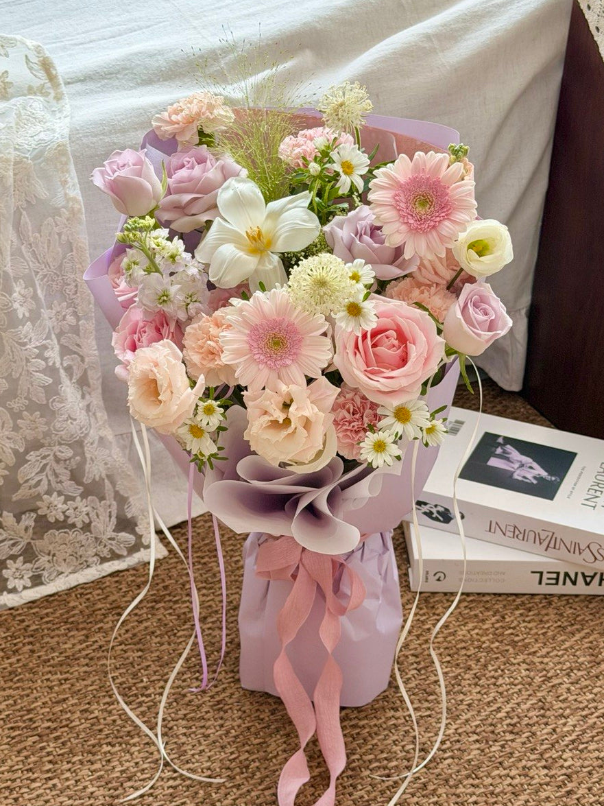 A pastel flower bouquet wrapped in light purple paper and ribbon, placed on a carpeted floor beside a bed with white bedding and a Chanel book.