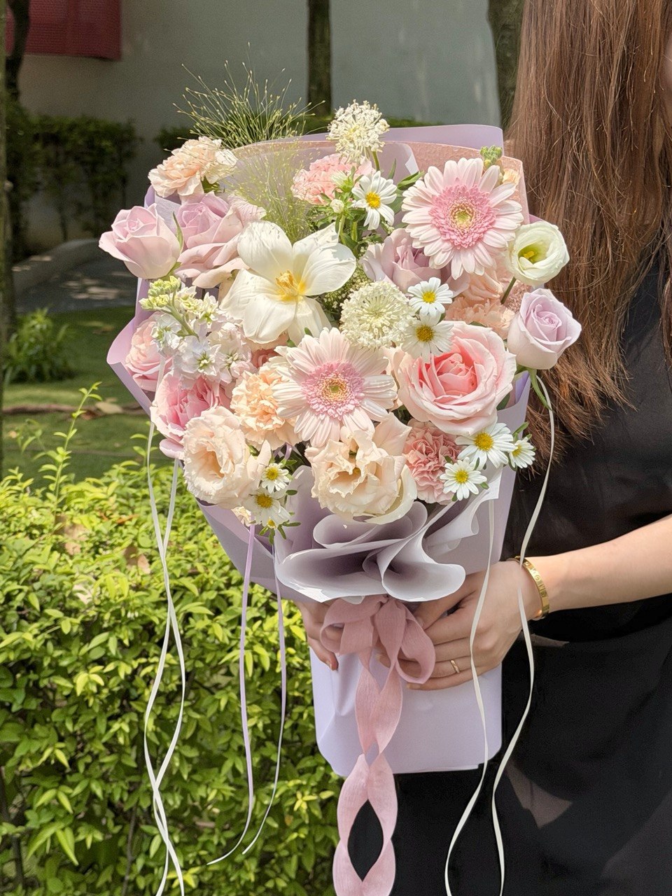 A pastel pink and cream flower bouquet held by a person outdoors. The bouquet features pink and white roses, daisies, and other soft pink blossoms wrapped in pale pink paper with long ribbon tails.