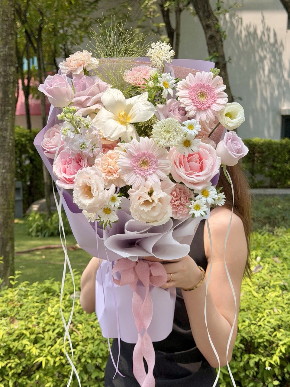 A person outdoors holding a large, vibrant bouquet of pink, peach, and white flowers wrapped in light purple paper with a pink ribbon.