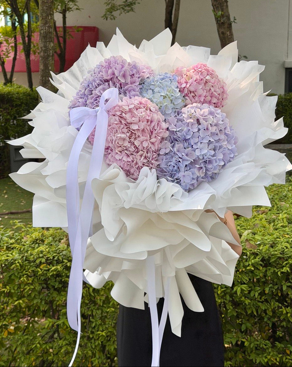 A large bouquet of pastel hydrangea flowers wrapped in white paper, held by a person outdoors in front of green hedges and trees. The flowers are in shades of pink, lavender, and light blue, with a pale purple ribbon tied around the bouquet.