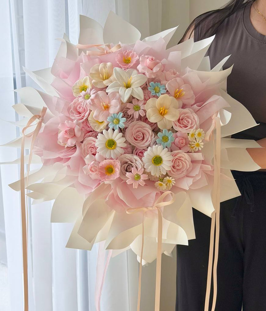 A large, beautifully wrapped bouquet of pink and white flowers with small blue and yellow accents, held by a person wearing a dark gray top and black pants in front of a bright window with white curtains.