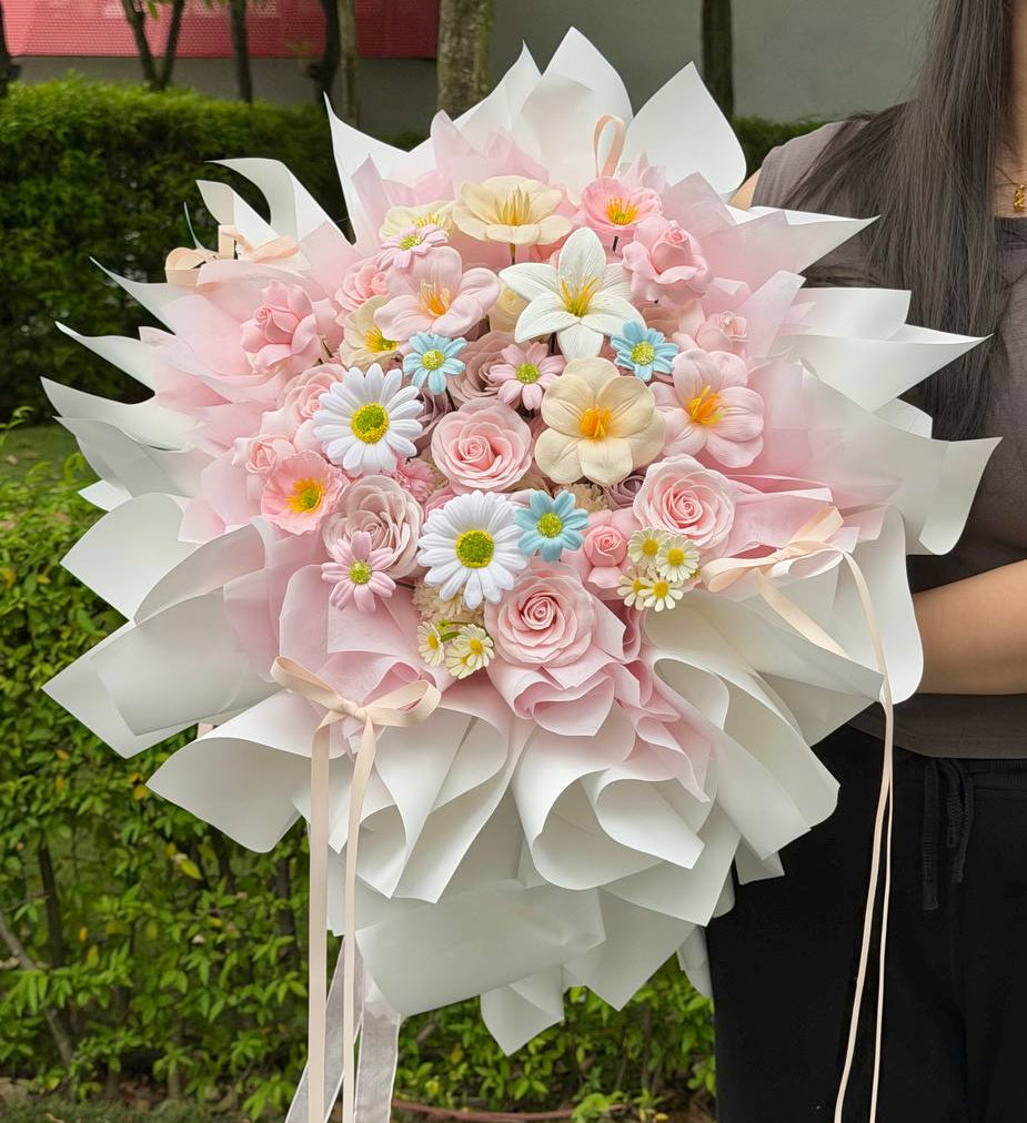 A large, beautifully arranged flower bouquet wrapped in white and light pink paper, held by a person outdoors in front of a garden background.