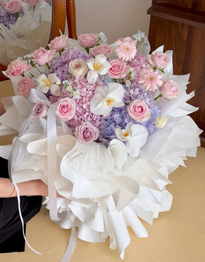 A large, beautifully arranged bouquet of pink, purple, and white hydrangea wrapped in white paper, held by a person whose arm is partially visible on the left, with a wooden cabinet in the background.