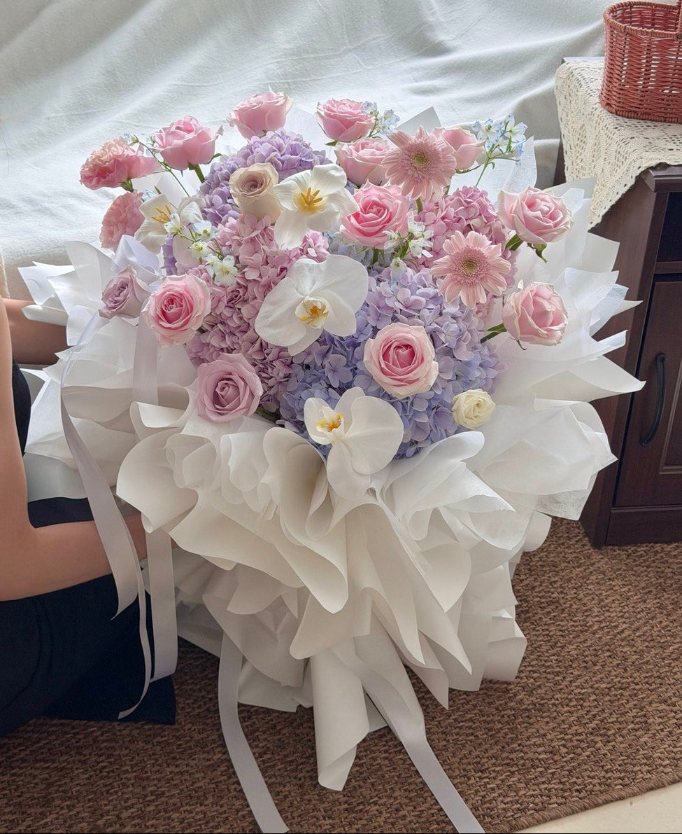 A large floral bouquet with pink roses, purple hydrangeas, white orchids, and pink daisies wrapped in white tissue paper, photographed indoors on a carpeted floor with a white sheet in the background.
