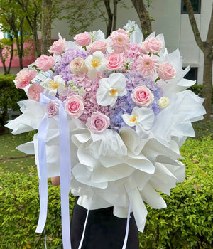 A large, round floral bouquet featuring pink and white roses, purple hydrangeas, and white orchids, wrapped in white tissue paper with long white ribbons, held outdoors in front of a garden background.