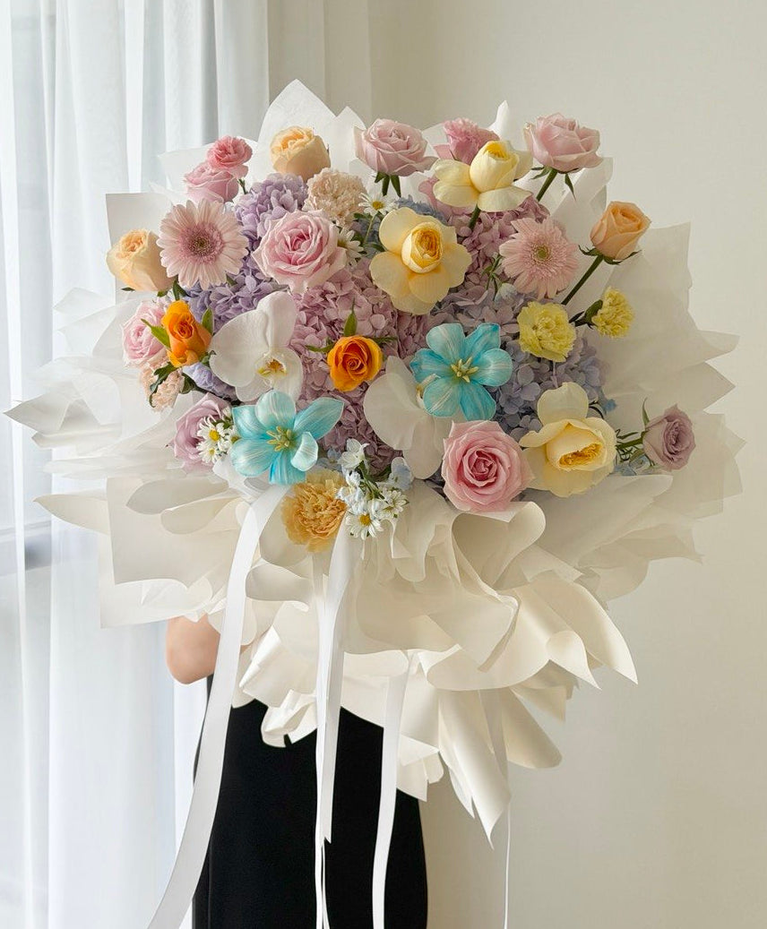 A large, vibrant bouquet of multicolored flowers wrapped in white wrapper and tied with white ribbons, held by a person wearing a black skirt in front of a white curtain.