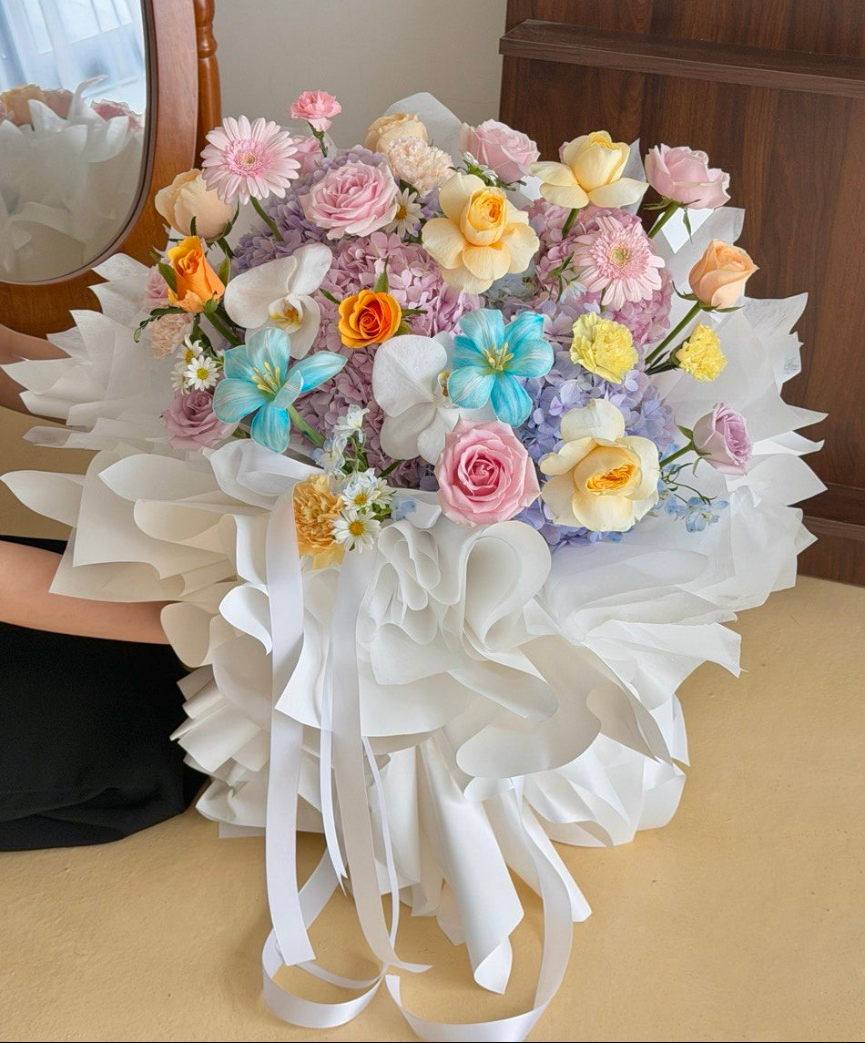A large, vibrant bouquet of multicolored flowers and hydrangea wrapped in white paper with long white ribbons, held by a person whose arm and hand are partially visible on the left, set against an indoor background.