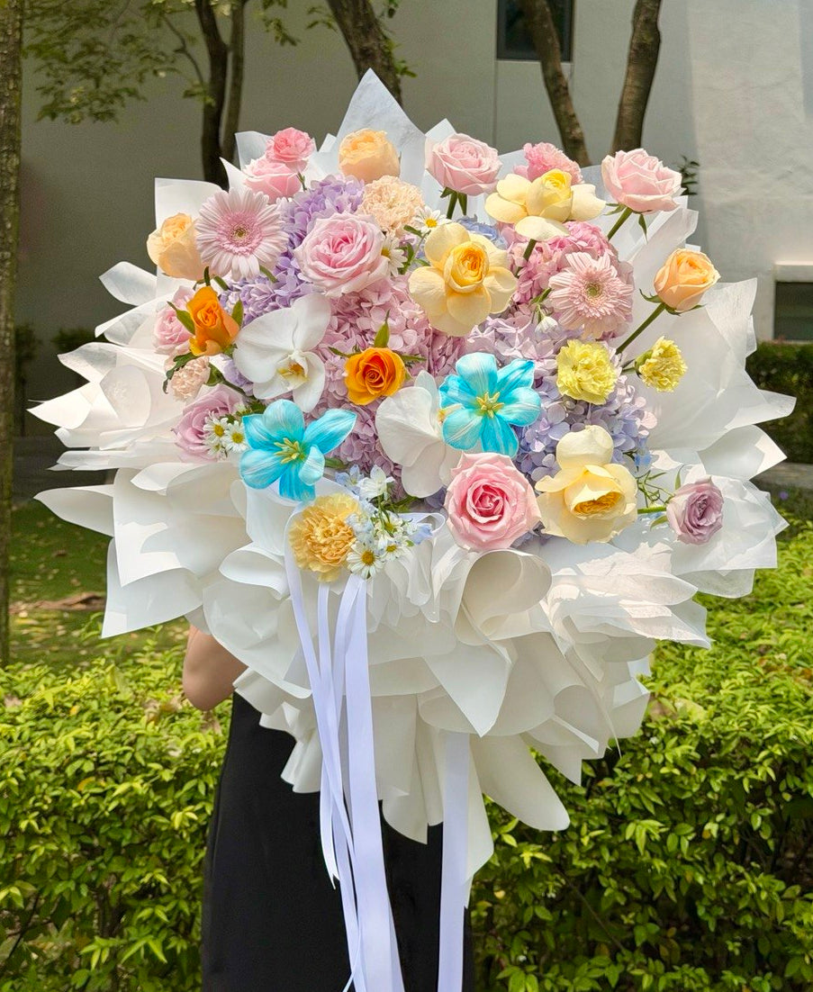 A large, vibrant bouquet hydrangea bouquet mixed with roses and tulip wrapped in white wrapper in ballet style with long white ribbons, held by a person outdoors in front of green bushes and trees.